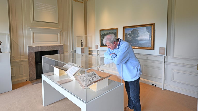 Man looking into a glass cabinet with large botanical books and oil painting behind him